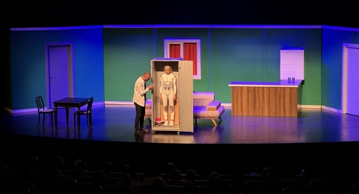 Theater scene: one man stands beside a glass cabinet containing another man on a stage with a living-room set (sofa, table) and a kitchen counter in the background.