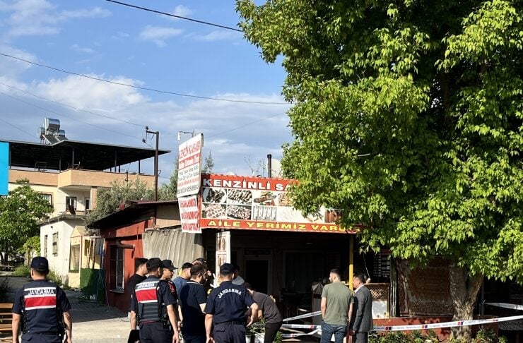 Group of uniformed police officers and civilians outside a small shop with red-and-white caution tape across the entrance on a sunny day.