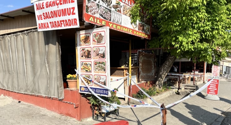 Exterior view of a Turkish family restaurant with an outdoor seating area, a large menu board showing dishes, and red-and-white banners; the entrance is cordoned off with blue-and-white caution tape.