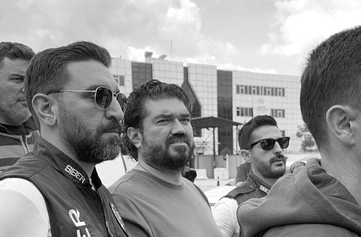 Group of men with beards and sunglasses standing together outdoors near a modern building, looking in the same direction.
