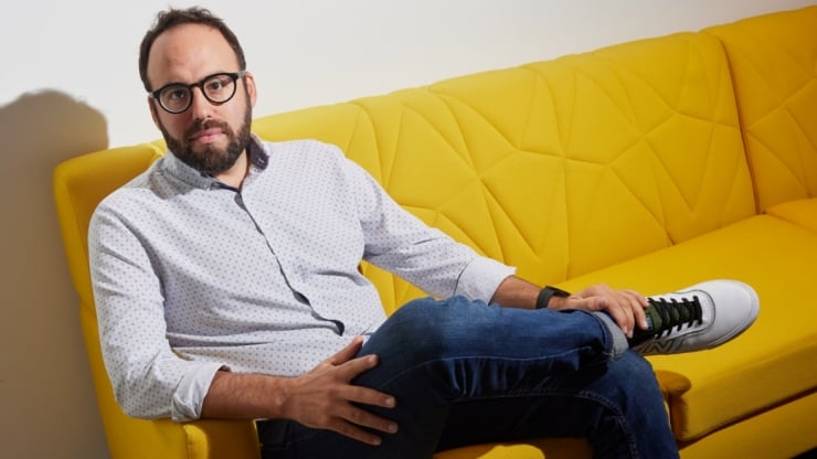 Man with glasses and beard sits casually on a bright yellow quilted sofa, legs crossed, hands resting on his knee.