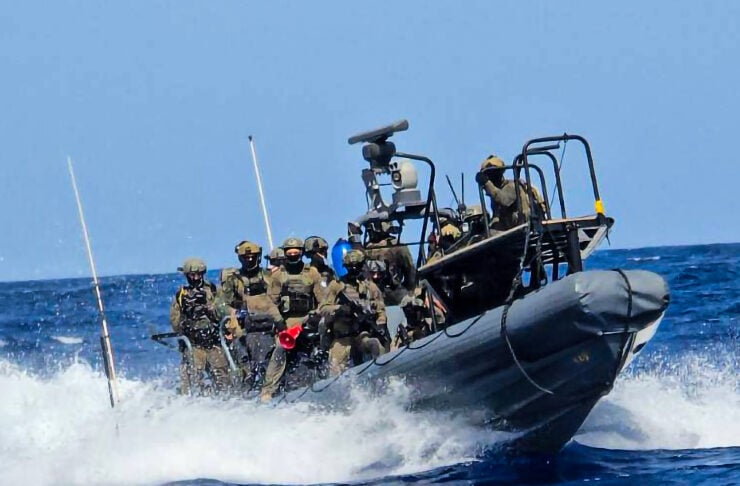 Military boat speeds through rough sea with armed personnel on board, waves splashing around.