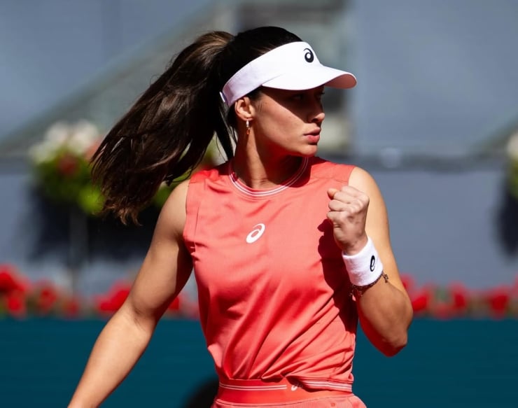 Female tennis player in a coral sleeveless top and white visor celebrates with a clenched fist on the court.