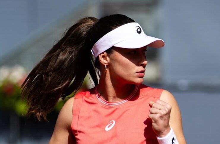 Female tennis player in a coral sleeveless top fists-pumping on a tennis court, wearing a white visor and wristband, hair in a ponytail.