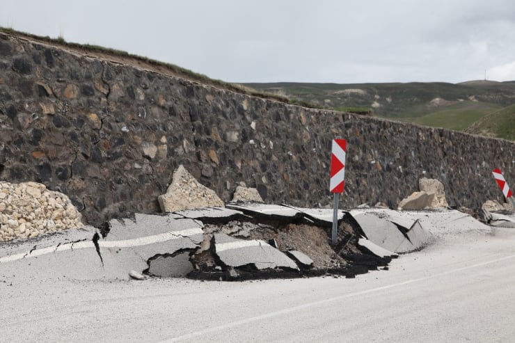 Collapsed road with cracked asphalt beside a stone retaining wall and red/white chevron hazard posts nearby.