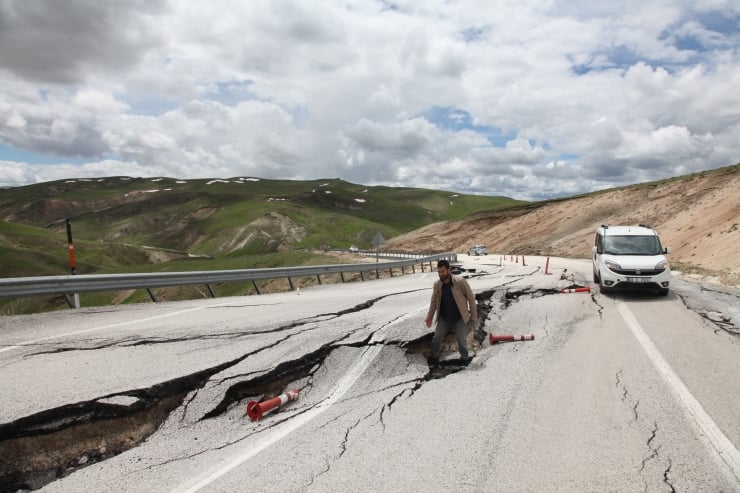 Worker walks on a cracked mountain road with a damaged lane, a white van nearby, and guardrails along a collapsed highway scene.