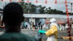 Silhouetted onlooker watches a healthcare worker in a yellow hazmat suit and white hood handling a garbage bag at a makeshift beachside aid camp with plastic chairs in the background, suggesting medical support or waste handling.