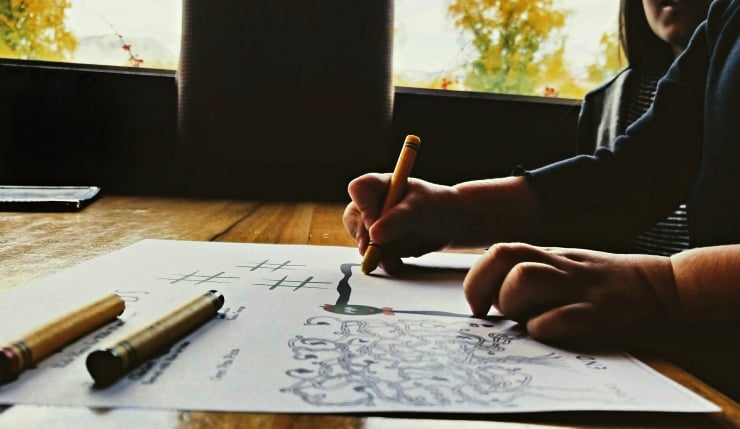 Child's hands drawing with a yellow marker on a paper filled with doodles and tally marks at a wooden table by a sunny window.