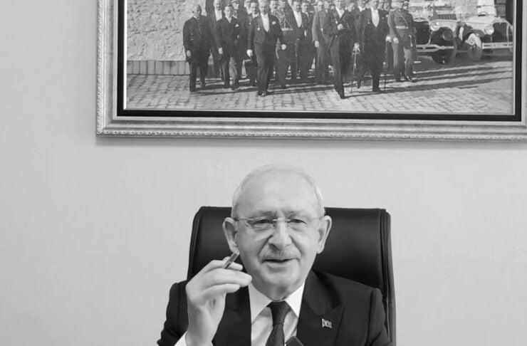 Older man in a suit sits at a desk with papers, holding a pen and smiling in a formal office setting.