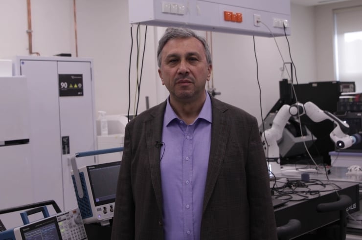 Man in a blazer stands in a laboratory with testing equipment and robotic arms in the background.