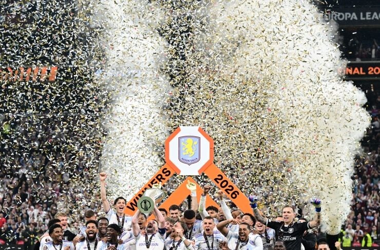 Soccer team in white celebrate on the pitch around a large trophy as gold confetti rains down, with a 'Winners 2026' banner in the background.