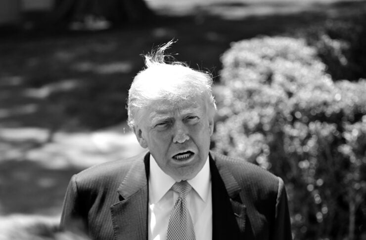 Man in a dark suit and light tie speaks to reporters outdoors, microphones in view in a black-and-white photo.