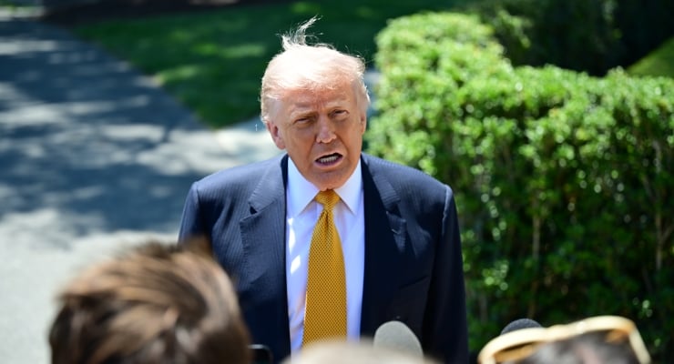 Man in a navy suit with a yellow tie speaks to reporters outdoors, microphones in front.