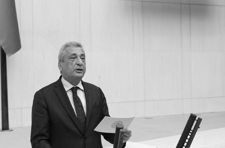 Older man in a dark suit delivering a speech at a podium, holding a document in one hand.