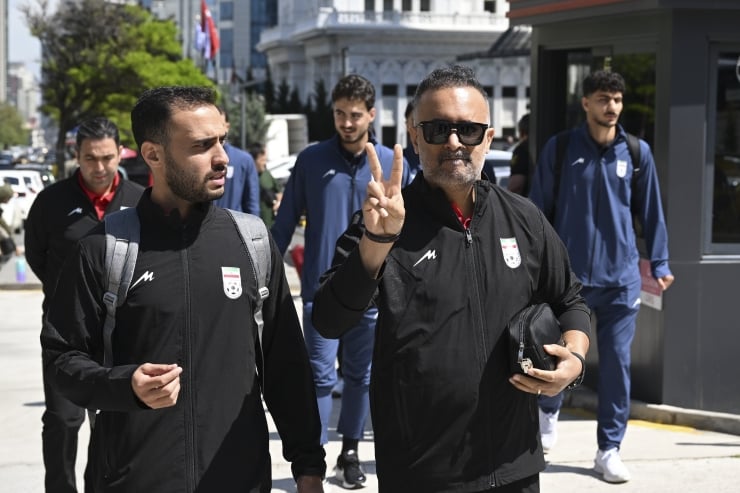 Group of men in athletic jackets walk on a sunny city street, one man in sunglasses holds up a peace sign to the camera.