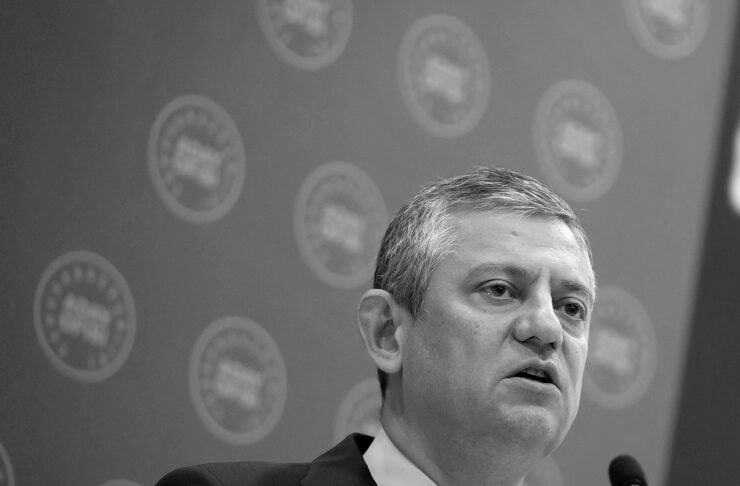 Man in a suit and tie speaking at a podium with microphones during a press conference, backdrop covered with circular logos.