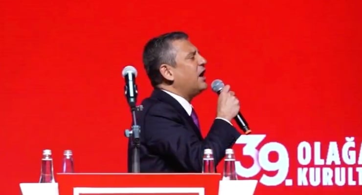 Male speaker in a dark suit and purple tie, holding a microphone at a red podium during a conference or event.