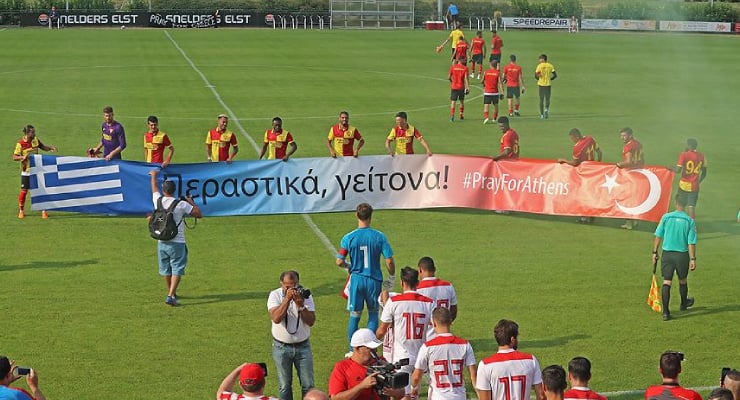 Football match scene with players holding a long banner featuring Greek and Turkish flags and the hashtag #PrayForAthens on a sunny field