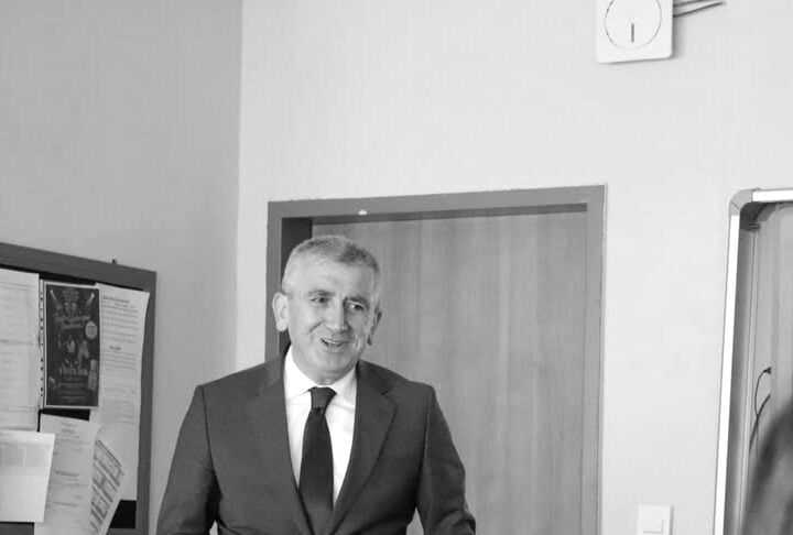 Smiling man in a dark suit and tie adjusts his jacket in a crisp office setting, with a door behind him and a bulletin board nearby.