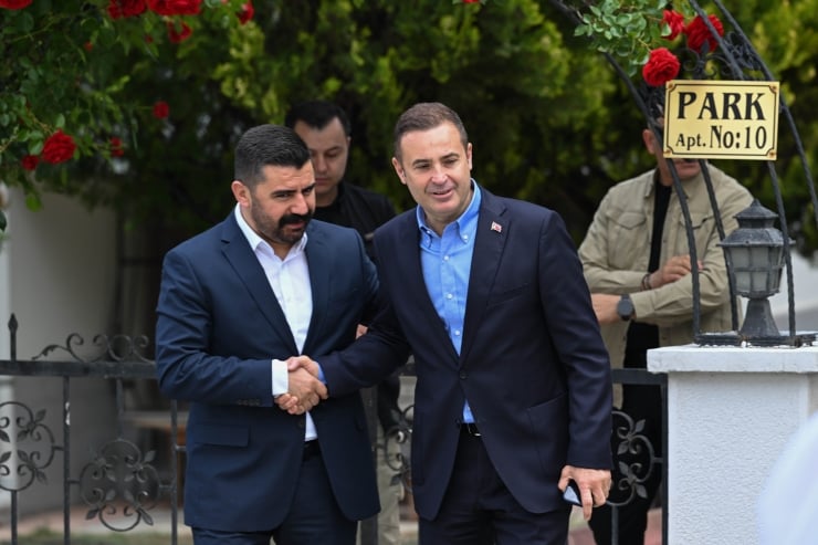 Two men in suits shake hands outside a building, smiling, with a decorative fence and a 'PARK Apt. No:10' sign nearby.