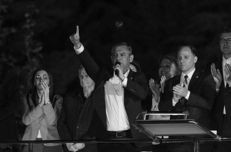 Speaker on a nighttime campaign stage, microphone in hand, raises one finger as supporters applaud behind him; banner with Turkish text visible in foreground.