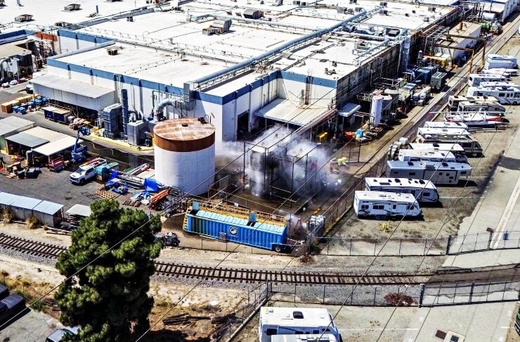 Aerial view of a large industrial facility with white buildings, a rusted cylindrical tank, fenced yard, and many RVs and trucks parked nearby.