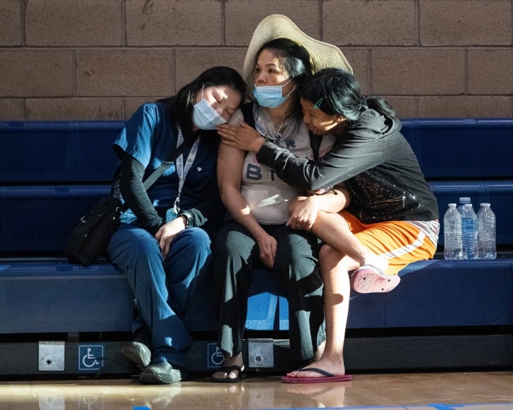Three people in masks sit on blue gym bleachers, two leaning on the middle person wearing a straw hat, with water bottles nearby.
