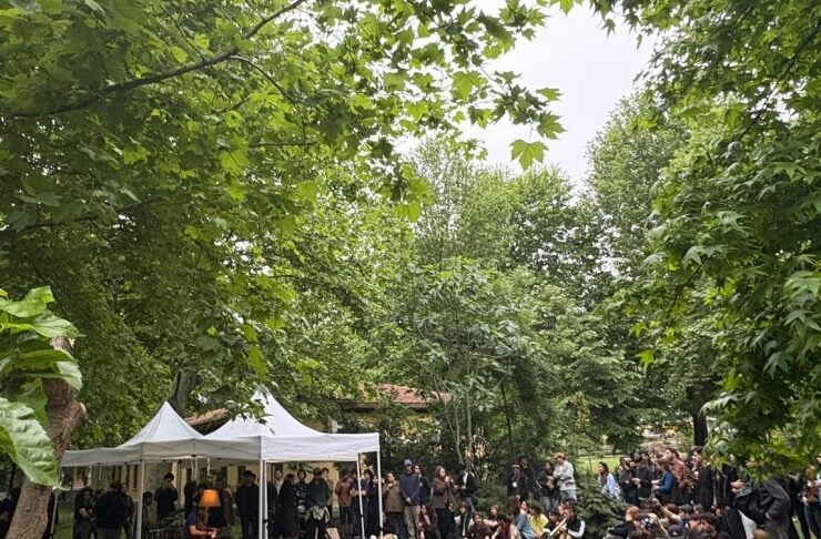 Outdoor gathering in a park with a white canopy tent and a crowd seated on the grass and benches, listening to a performer on stage left.