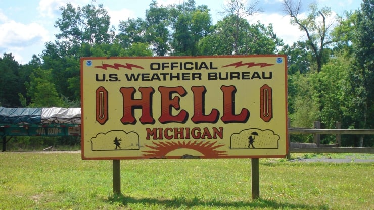 Yellow roadside sign announcing 'HELL, MICHIGAN' with U.S. Weather Bureau header and sun graphic, trees in background.