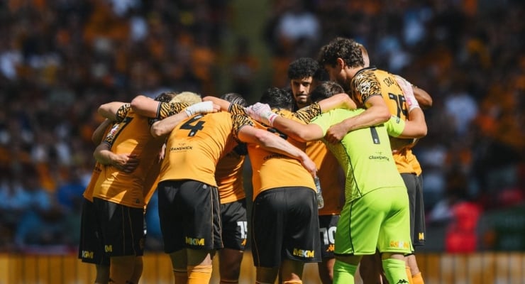 Soccer team in yellow jerseys huddles in a circle on the field before a match, goalkeeper in neon green mitts.