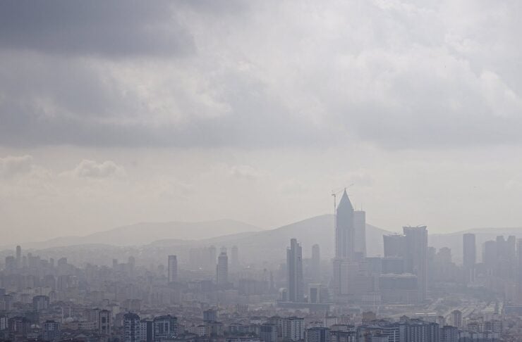 City skyline with tall buildings hazy in the air, distant mountains, and trees in the foreground under a cloudy sky.