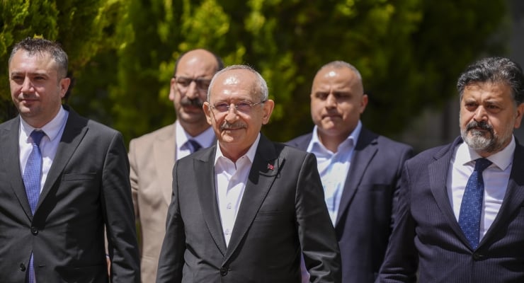 Five men in suits walking outdoors on a sunny day, with a central man wearing glasses and a mustache leading the group.