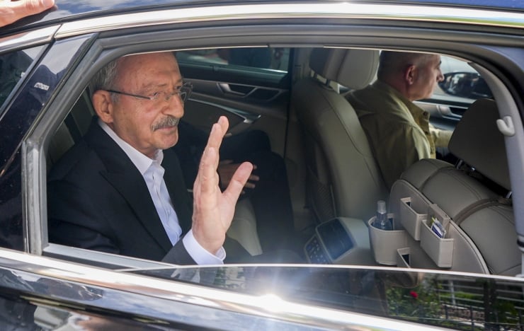 Older man in a suit in the back seat of a car, waving hello as a driver sits in front.