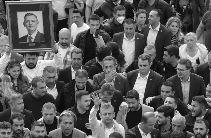 Large black-and-white crowd of people at an outdoor event, with one person holding a framed portrait of a man above the crowd.