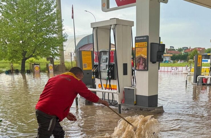 Man in a red shirt wading through floodwater at a Shell gas station, using a pole to stir muddy water near pumps.