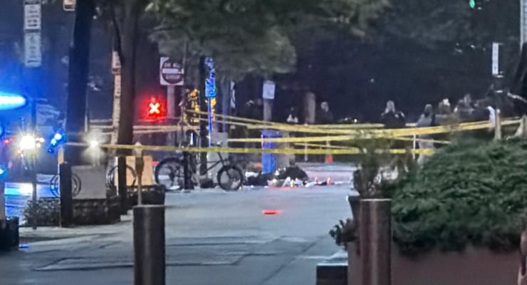 Nighttime street scene with yellow police tape cordoning off a sidewalk, a person lying on the pavement near the tape, bicycles nearby, and blue emergency lights from a police vehicle in the background.
