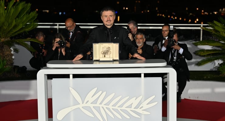 A man stands behind a modern lectern on a red-carpeted stage at night, holding an award while photographers film from behind him.