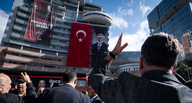 Crowd of people at a political rally outside a glass-front building, with large banners including the Turkish flag and a portrait of a man.
