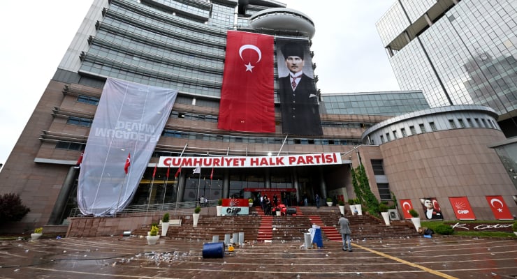 Exterior of a modern building draped with a large Turkish flag, a portrait banner, and a CHP banner reading 'Cumhuriyet Halk Partisi' above the entrance; people on the steps.