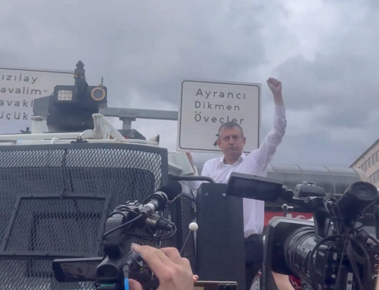 Man in a white shirt on an armored vehicle raises his fist as cameras film in the foreground.