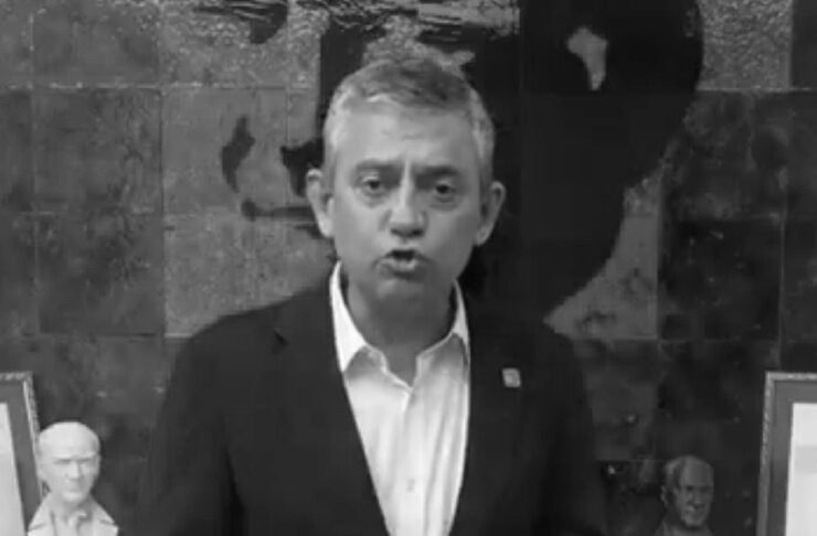 Man in a dark suit speaks to the camera with hands clasped, standing in front of framed busts and a dark wall backdrop (black and white).
