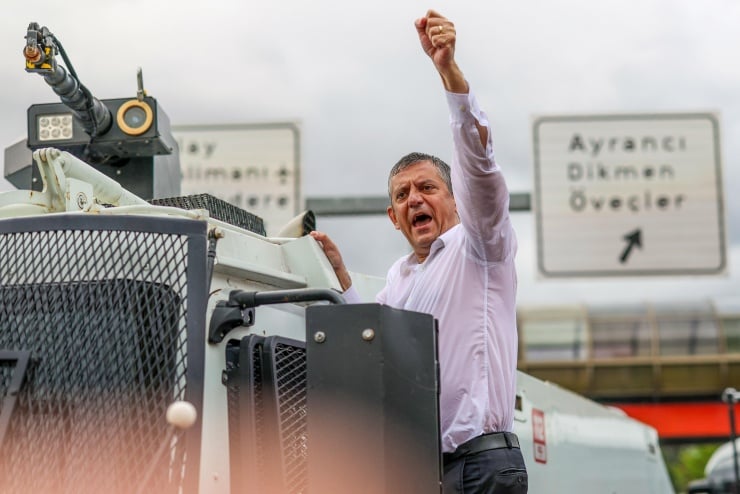 Man in a white shirt stands on a large vehicle, fist raised in the air during a protest on a highway with road signs in the background.