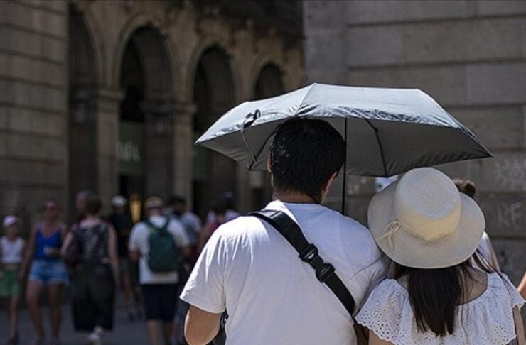 Couple walking arm in arm under a shared umbrella in a sunny city street, with pedestrians in the background.