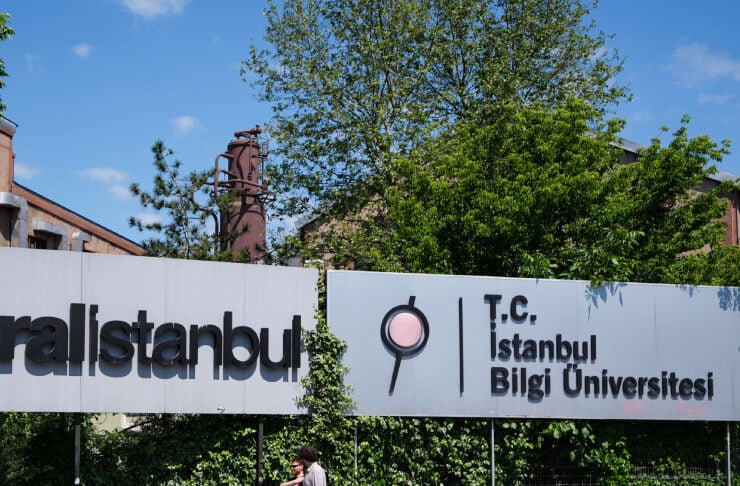Exterior sign reading 'T.C. Istanbul Bilgi Üniversitesi' with a circular logo, on a sunny day with greenery in the background, and two people walking on the sidewalk nearby