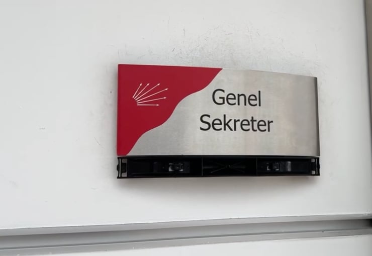 Metal sign on a white wall reading 'Genel Sekreter' with a red curved design and a small logo on the left.