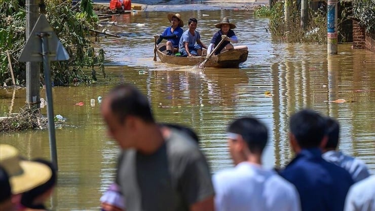 People in a small wooden boat navigate murky floodwaters along a flooded street; blurred figures in the foreground watch the scene.