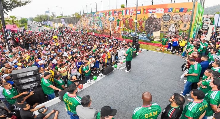 Large crowd gathered at an outdoor event, with a speaker on a stage facing fans wearing green jerseys, and a colorful mural wall in the background.