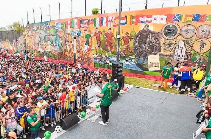 Speaker in a green shirt addresses a large outdoor crowd from a small stage, with a mural-covered wall in the background.