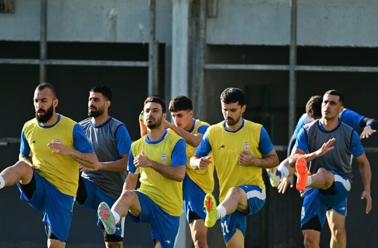 Soccer players in yellow and blue jerseys perform high-knee drills on a grass field during practice, with cones and a ball nearby.