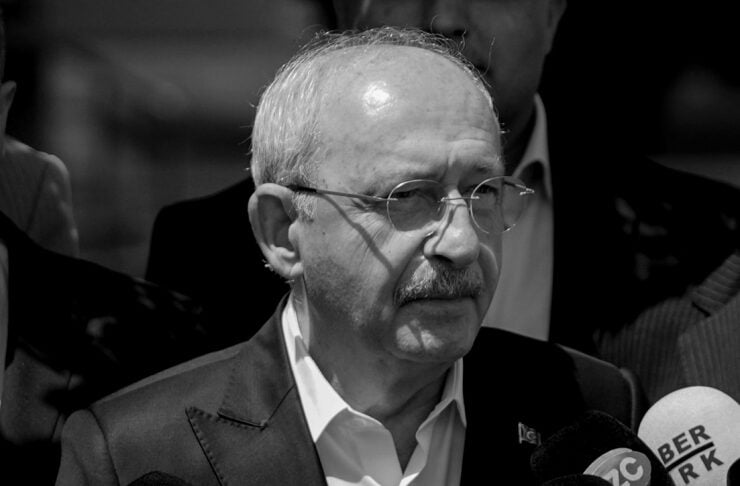 Older man in a suit speaks to reporters, surrounded by numerous microphones from various news outlets